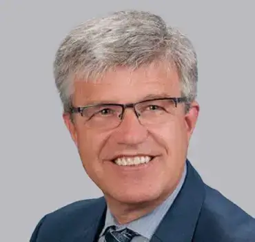 Professional man with gray hair and glasses, smiling in a business suit against a gray background.