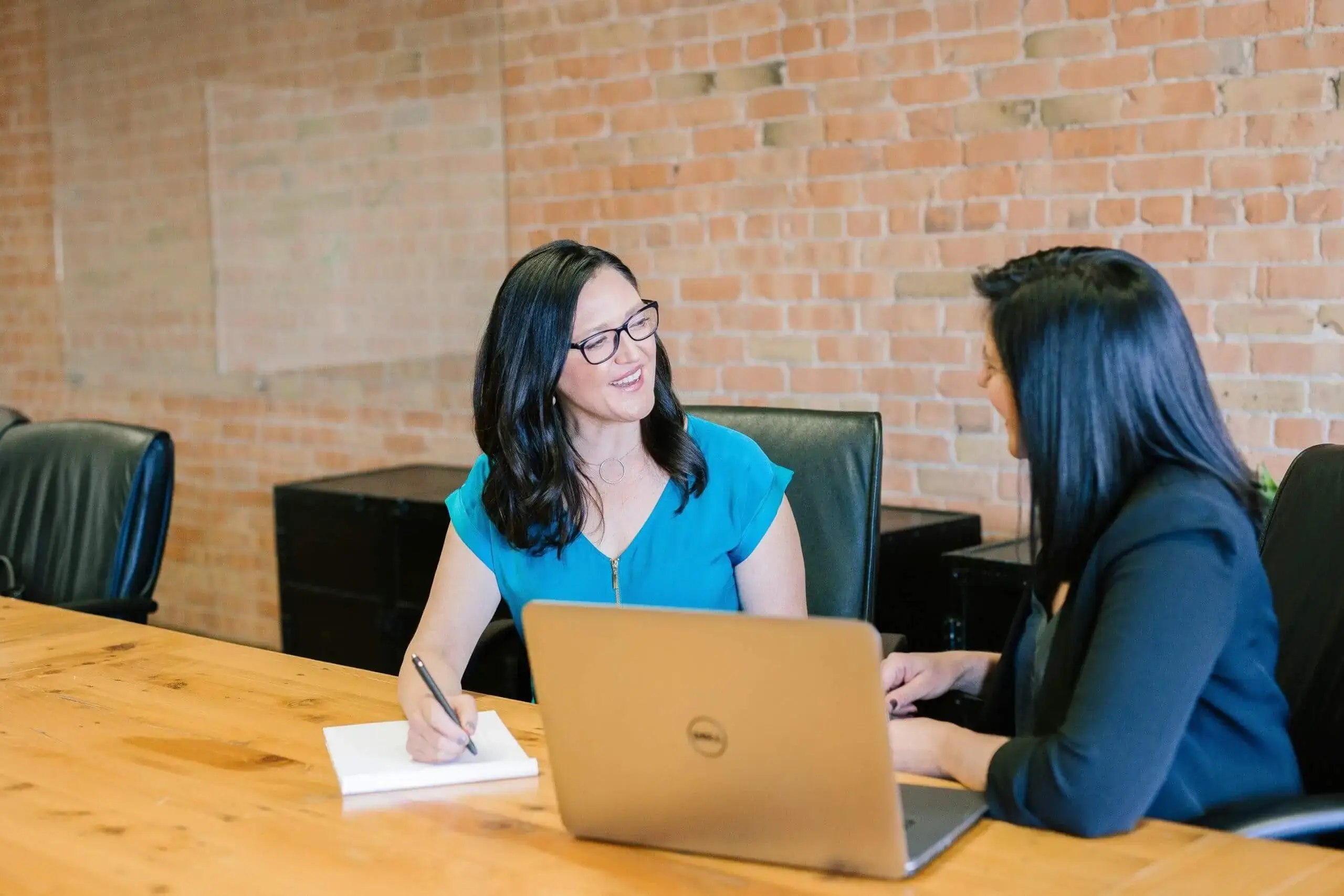 Two professional women engage in a collaborative discussion at a conference table, with a laptop and notes in view.